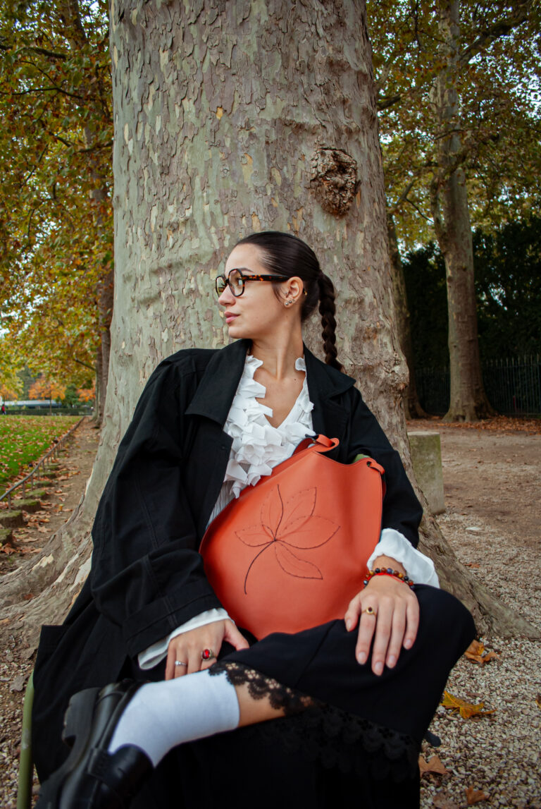 Photo de mode avec une mannequin appuyée contre un arbre portant un manteau noir et un sac à main rouge