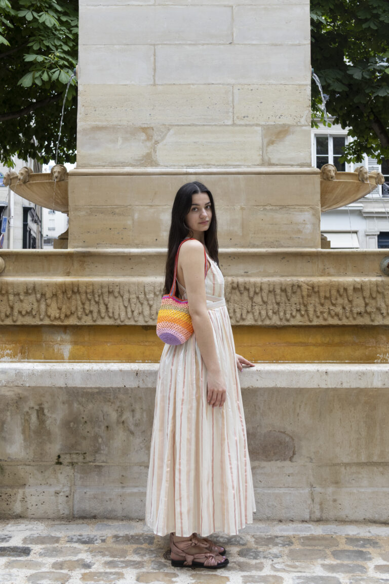 Photo de mode avec une mannequin de profil appuyée sur une fontaine avec une robe en tissu et portant un petit sac coloré.